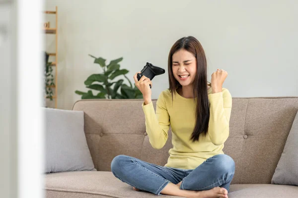 若いアジアの女性は自宅のリビングルームで大きな快適なソファに座っている間にゲームに勝つと楽しさと両手を上げてゲームをプレイするためにビデオゲームコントロールジョイスティックを使用しています — ストック写真