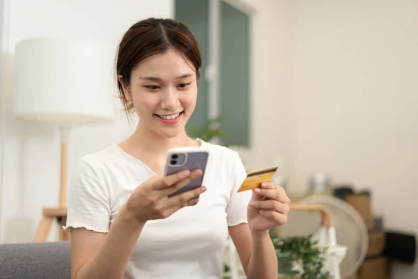 Teenage girl is holding credit card and typing credit card number for payment after shopping online on smartphone while sitting to relax comfortable on the couch in living room.