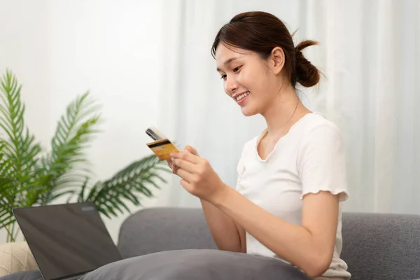 Teenage girl is holding credit card and typing credit card number for payment after shopping online on smartphone while sitting to relax comfortable on the couch in living room.