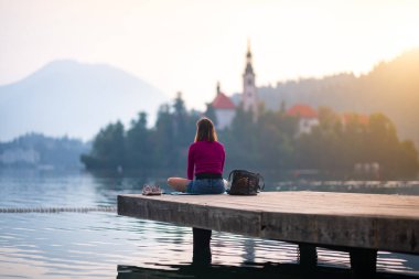 Tahta bir iskelede yalnız bir kız oturuyor. Arka planda güzel manzarası olan meditasyon yapan bir kadınla sakin bir sahne..