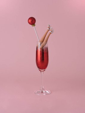 Bebeklerin bacakları, pastel pembe arka planda kırmızı parıltılarla dolu bir martini bardağında banyo yapıyor. Yaratıcı asgari parti konsepti. Yeni yıl ve mutlu noeller ilham kaynağı.