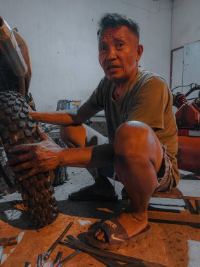 an old man in short shorts is trying hard to change the rear motorcycle tire at night in his workshop.  photo taken at night outdoors in luhu village, gorontalo on august 21, 2022
