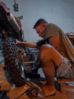 an old man in short shorts is trying hard to change the rear motorcycle tire at night in his workshop.  photo taken at night outdoors in luhu village, gorontalo on august 21, 2022