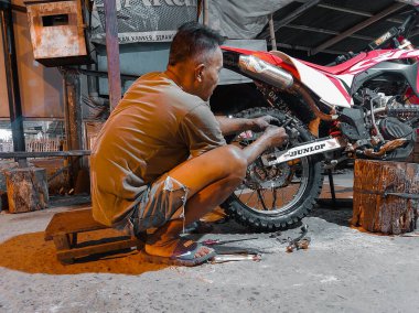an old man in short shorts is trying hard to change the rear motorcycle tire at night in his workshop.  photo taken at night outdoors in luhu village, gorontalo on august 21, 2022