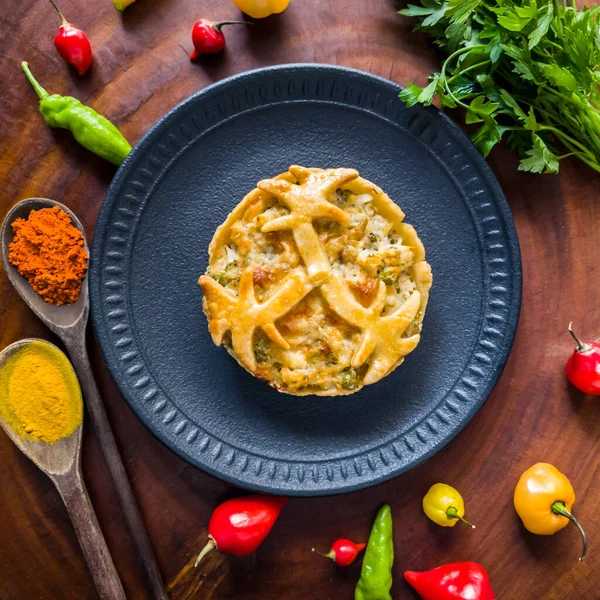 Tasty small chicken pie on black plate with peppers, wooden spoon parsley, saffron and spicy paprika on wooden background. Top view of chicken pie with spices and vegetables.