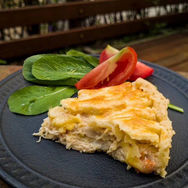Slice of tasty chicken pie with arugula and tomatoes on black dish and wooden background. Close-up, side view.