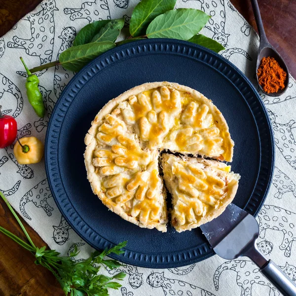 tasty chicken pie on black plate with yellow and red peppers, green leaves, wooden spoon, parsley and paprika on wooden background. Top view of pie slice with spices and vegetables.