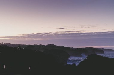Güneş okyanusun üzerinde batarken, gökyüzünü mora boyarken, ön plandaki kayalar ufka karşı güzel bir doğal manzara yaratır.
