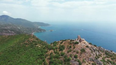 Dağın tepesindeki antik ve ortaçağ binasının havadan görünüşü, yeşil bitki örtülü kayalık kıyı şeridi, aşağı mavi deniz. Beyaz kayalar, ağaçlar. Dalgalar boş bir plaja çarpıyor, mavi gökyüzü bir yaz günü..