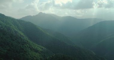 Yazın el değmemiş yeşil dağın havadan görünüşü. Güneş ışığı bulutların üzerinden geçer. Arka planda sisli sıradağlar var. yavaş çekim