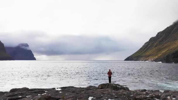 Vista aérea de mujer irreconocible se para en acantilado de roca mirando el fondo salvaje de las islas Fero.Belleza naturaleza prístina, montañas verdes.Video de mujer admirando las olas rompen en acantilados de rocas — Vídeos de Stock