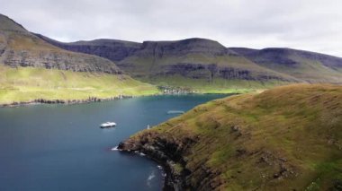 Faroe adalarındaki ünlü göl manzarası, göldeki tekneler. Rocky sahili, dalgalar, sıradağlar, yeşil çayır ve bulutlu gökyüzü. Mavi gölü ve arka planda küçük bir köyü olan dağ manzarası.