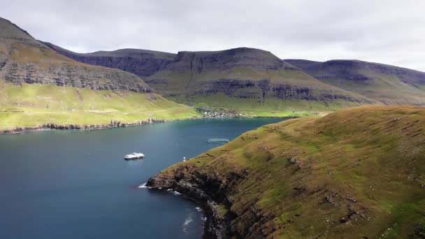 Flygfoto över berömda sjön i Färöarna, båtar i sjön. Klippig kust, vågor, bergskedja, grön äng och molnig himmel. Bergslandskap med blå sjö och liten by i bakgrunden. — Stockvideo