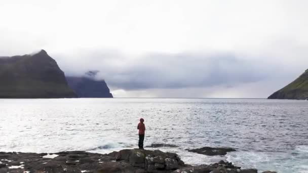 Et flybilde av en ugjenkjennelig kvinne står på en klippe med utsikt mot villmarksbakgrunn av Færøyene. Skjønnhet, ugjenkjennelig natur, grønne fjell. Video av kvinner som beundrer bølger som knuses på klipper – stockvideo