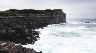 Hava manzaralı yüksek kaya kayalıkları, okyanus dalgaları kayalıklara çarpıyor. Okyanusun gücü, güçlü dalgalar köpük yaratıyor, vahşi doğa, vahşi yaşam, vahşi insanlar yok, gökyüzünde bulutlar, manzaralar