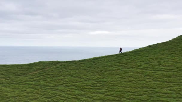 Vista aérea de mulher irreconhecível andando em uma estrada suja em um cume de montanha verde com mochila. Atividades desportivas e atlético e desportivo pessoa caminhando para o farol, nuvens no céu. — Vídeo de Stock