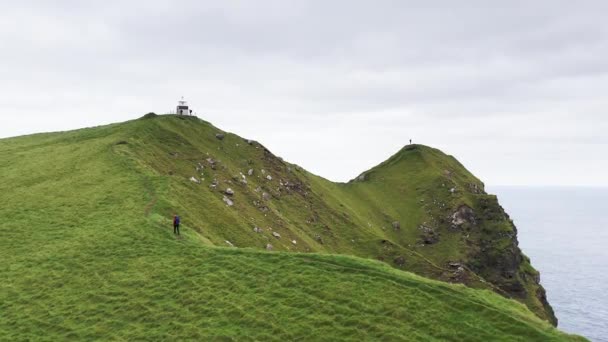 Vista aérea de una mujer irreconocible caminando por un camino sucio en una cresta de montaña verde con mochila. Actividades deportivas y deportistas caminando hacia el faro, nubes en el cielo. — Vídeos de Stock