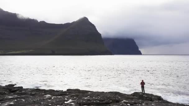 Vista aérea de mujer irreconocible se para en acantilado de roca mirando el fondo salvaje de las islas Fero.Belleza naturaleza prístina, montañas verdes.Video de mujer admirando las olas rompen en acantilados de rocas — Vídeos de Stock