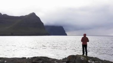 Faroe Adaları 'nın vahşi arka planına bakan tanınmayan bir kadının havadan görünüşü. Güzel, el değmemiş doğa, yeşil dağlar. Kayalıklardaki dalgaları hayranlıkla seyreden bir kadının videosu.