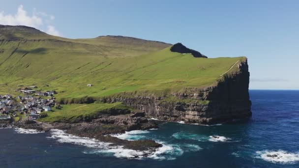Dron přelétající nad mořem, obklopený obrovským a majestátním útesem, pokrytým zelenou trávou. Malá vesnička v suterénu útesu v letním slunečném dni. — Stock video