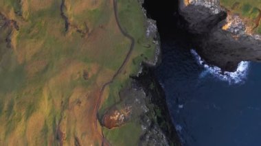 Faroe Adaları 'nın büyük uçurumlarının üst hava manzarası. Gün batımında bir V koyunun üzerinde uçan insansız hava aracı. Taş kayalıklarda yeşil ve kahverengi çayırlar. Uçurumun kenarındaki dalgalı mavi deniz
