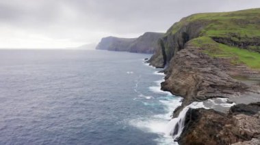 Faroe Adaları 'ndaki kaya uçurumunun havadan görünüşü. Mavi deniz dalgaları, bulutlu yolculukta dalgalı deniz. yavaş çekim