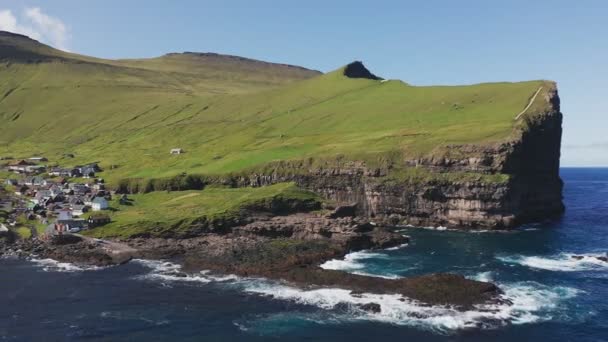Drone volant au-dessus de la mer, encadrant une énorme et majestueuse falaise, couverte d'herbe verte. Petit village au sous-sol de la falaise par une journée ensoleillée d'été. — Video