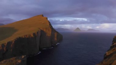 Gün batımında altın arazinin havadan görünüşü. Faroe Adası 'ndaki uçurumun arka planı, mavi deniz ve adalar insansız hava aracı videosunda.