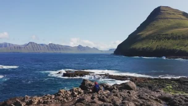 Drone volant au-dessus de la plage de rochers, l'homme assis devant le paysage. Jeune homme se reposant et regardant mer bleue rugueuse, île verte et falaise en arrière-plan pendant la journée ensoleillée. — Video