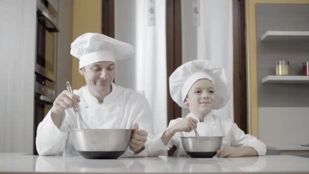 Táta a syn míchají jídlo v ocelové misce ve své kuchyni. Okamžiky otce a syna při vaření. Otec učí syna vařit doma dort. — Stock video