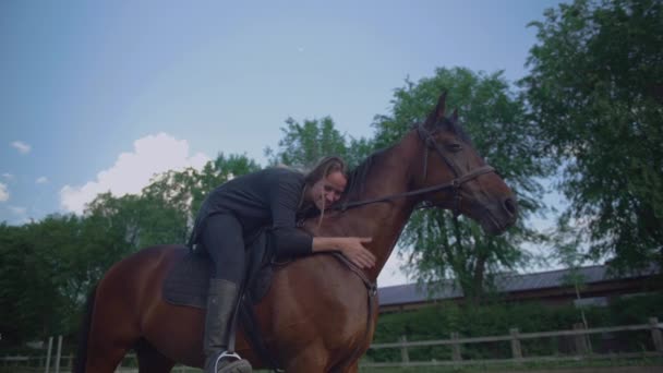 Jovem mulher sentada e abraçando cavalo em uma escola de equitação. Dia ensolarado. Cavalo entediado em pé em uma cerca — Vídeo de Stock