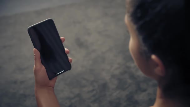 Vista posterior de la mujer fitness utilizando el teléfono inteligente en casa. Mujer que se prepara para el entrenamiento usando el teléfono inteligente.Mujer que hace un toque en el teléfono móvil mientras descansa durante el entrenamiento físico — Vídeos de Stock