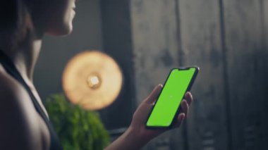 Evde yeşil ekran akıllı telefon kullanarak gülümseyen genç bir kadının yan görüntüsü. Kadın akıllı telefon kullanarak antrenmana hazırlanıyor. Kadın spor yaparken cep telefonu kullanıyor.