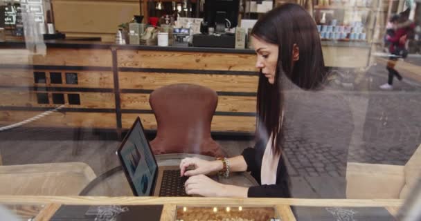 Zijaanzicht van een blanke vrouw die in een koffieshop werkt. Vrouw in slim casual pak schrijft over nieuw project op digitale laptop. Werk op afstand. Beelden gemaakt door het raam — Stockvideo