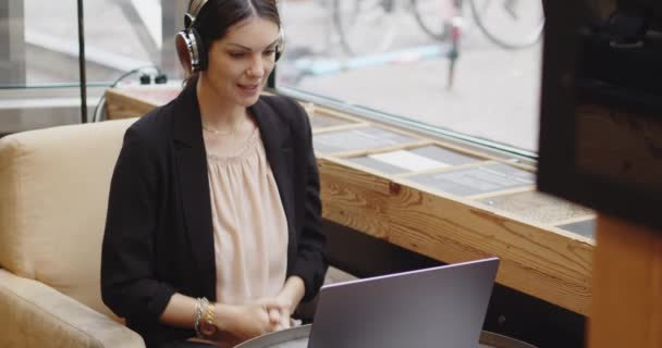 Νεαρή Καυκάσια γυναίκα με ακουστικά κατά τη διάρκεια συνάντησης σε δημόσιο μπαρ. Χαρούμενη νεαρή επιχειρηματίας σε casual κοστούμι καλώντας σε μια συνάντηση συνέδριο στην καφετέρια χρησιμοποιώντας σύνδεση στο internet. — Αρχείο Βίντεο