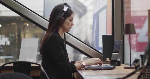 Seitenansicht einer kaukasischen Frau, die in einem Café arbeitet. Frau schreibt über neues Projekt auf digitalem Laptop und hört Musik mit ihren Kopfhörern. Arbeitsplätze aus der Ferne. Verfilmung durch das Fenster — Stockvideo