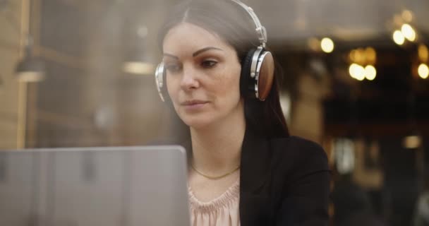 Blanke vrouw die in een koffieshop werkt. Vrouw luisterend en schrijvend in een webconferentie op de laptop, luisterend met een koptelefoon. Werk op afstand. Beelden gemaakt door het raam — Stockvideo