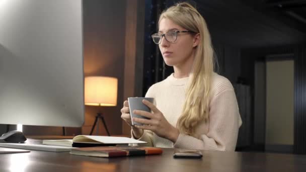 Νεαρή γυναίκα που ψάχνει υπολογιστή στο γραφείο. Γυναίκα που εργάζονται, πίνοντας φλιτζάνι καφέ κατά τη διάρκεια του βραδινού στο σπίτι — Αρχείο Βίντεο