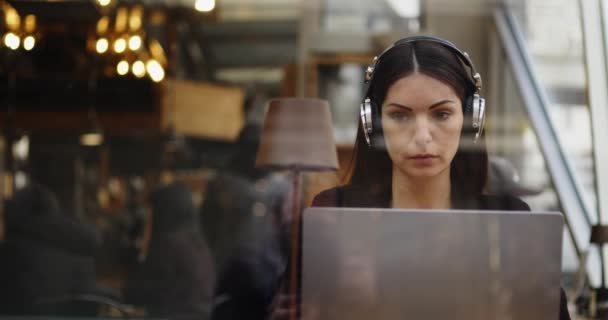 Mujer caucásica trabajando en una cafetería. Mujer escuchando y escribiendo en una conferencia web en el portátil, escuchando con auriculares. Trabajos de trabajo remoto. Filmación hecha por la ventana — Vídeos de Stock