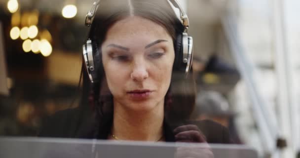 Kaukasische Frau, die in einem Café arbeitet. Frau im Gespräch hört Webkonferenz auf dem Laptop, hört mit Kopfhörern zu. Arbeitsplätze aus der Ferne. Verfilmung durch das Fenster — Stockvideo