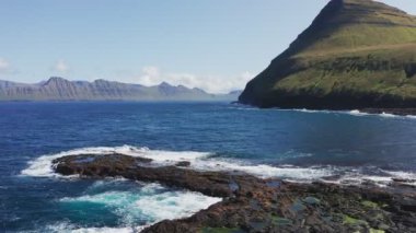 Kaya sahillerinde uçan drone, su birikintileri. Dalgalı mavi deniz manzarası, çimenli yeşil ada, dağın arka planı, güneşli bir günde uçurum. 