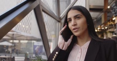 Gülümseyen genç bir iş kadını pencerenin önünde telefonla konuşuyor, mutlu bir iş kadını internet konferansını onaylamak için mobil arama yapıyor ve uzaktaki modern bir kafede duruyor. 4k