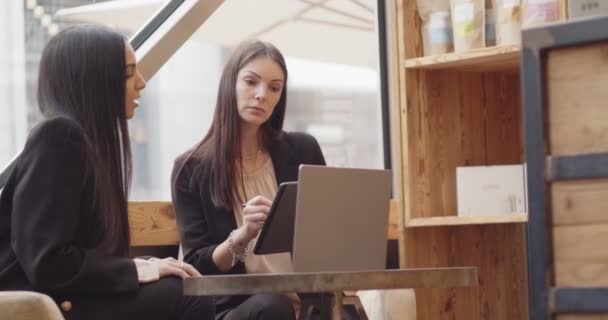 两名妇女坐在一家现代工业风格的咖啡馆里工作 两个在笔记本电脑和平板电脑上工作的多种族同事在聊天 团队合作 多样性和社会观念 — 图库视频影像