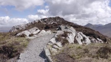 Güneşli bir günde, yazın bulutlarla birlikte İrlanda 'da taş patika boyunca panoramik bir yürüyüş. Vadinin aşağısındaki köy, arka planda deniz ve dağ sıraları. Kuru ve kurak çayır, çimen, kayalar. 4k