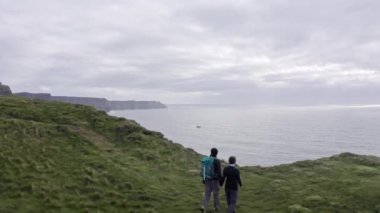 Aerial view of beautiful irish cliffs, young couple walk, watch the landscape, panorama hug each other. Cloudy sky in the background, rough sea. Famous place in Ireland, Mother of cliffs . 4k