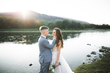 Zarif, şık, mutlu, sarışın gelin ve dağlardaki güzel bir nehrin arka planında harika bir damat.