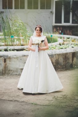 Zarif beyaz elbiseli güzel esmer gelin elinde buket ile güzel ağaçlar poz veriyor.