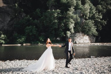 Zarif, şık, mutlu, sarışın gelin ve dağlardaki güzel bir nehrin arka planında harika bir damat..