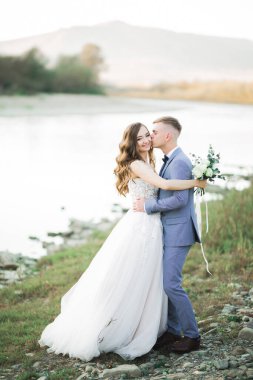 Güzel düğün çifti bir dağ nehrinin kıyısında taşlarla öpüşüp kucaklaşıyorlar..