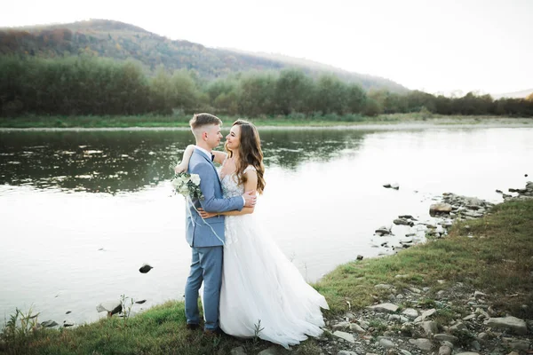 山の美しい川の背景にエレガントなスタイリッシュな幸せなブロンドの花嫁と豪華な新郎 — ストック写真